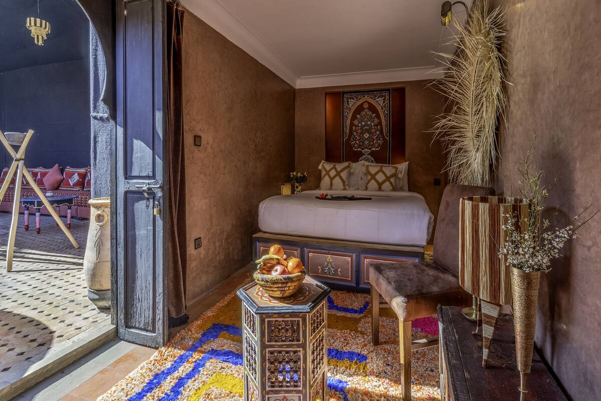 A cozy bedroom with a bed, a small table with a basket of fruits, a bench, and a decorative wall piece.