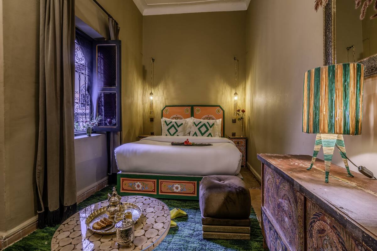 A hotel room with a bed, a nightstand, a table with a tray of food, and a decorative lamp.