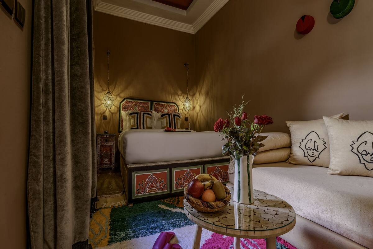 A bedroom with a bed, a bedside table with a vase and fruit basket, and a colorful rug.