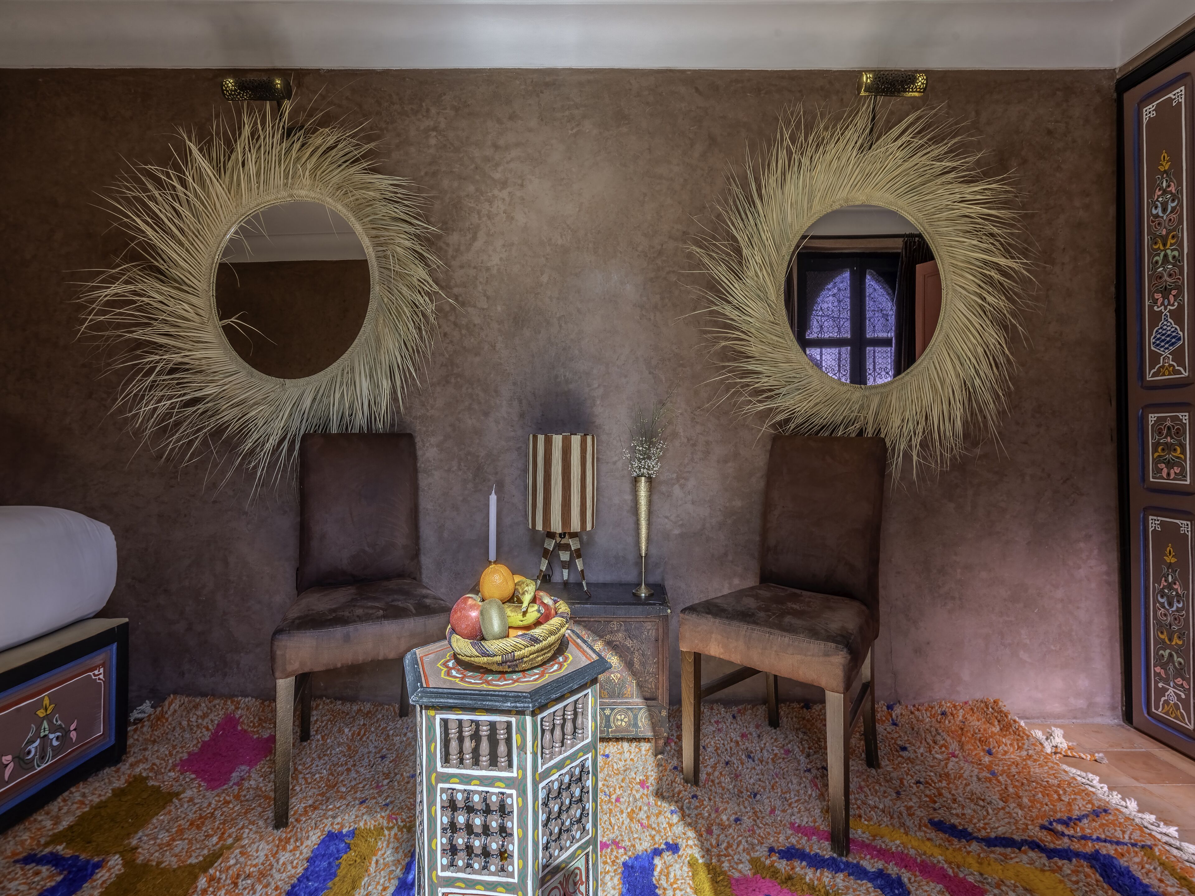 A room with two chairs, a small table with fruit, a decorative mirror, and a patterned rug.