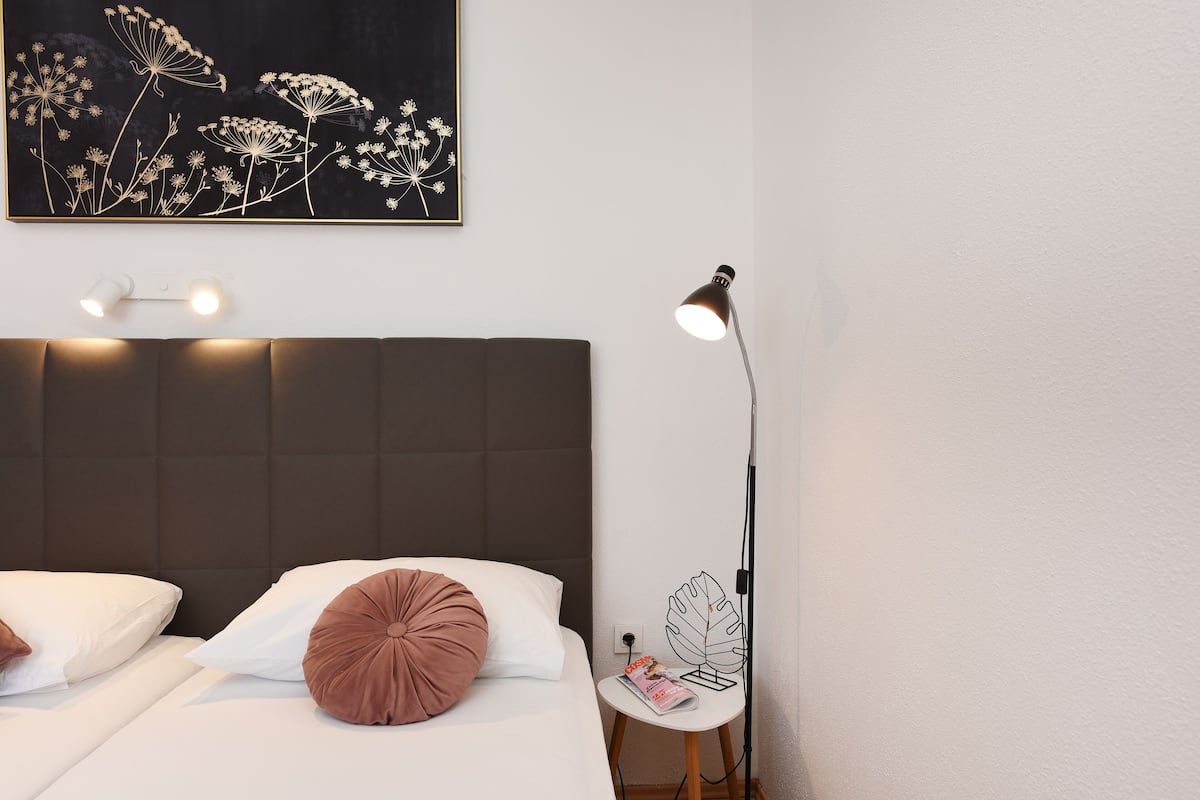 A neatly made bed with white and brown pillows, a dark headboard, a wall-mounted light fixture, and a floor lamp with a leaf-shaped base.