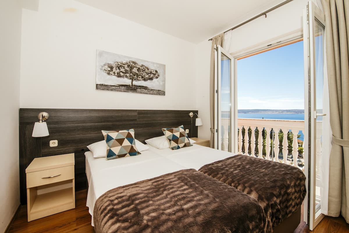 A bedroom with a large bed, a nightstand, a lamp, a painting of a tree, and a balcony with a view.