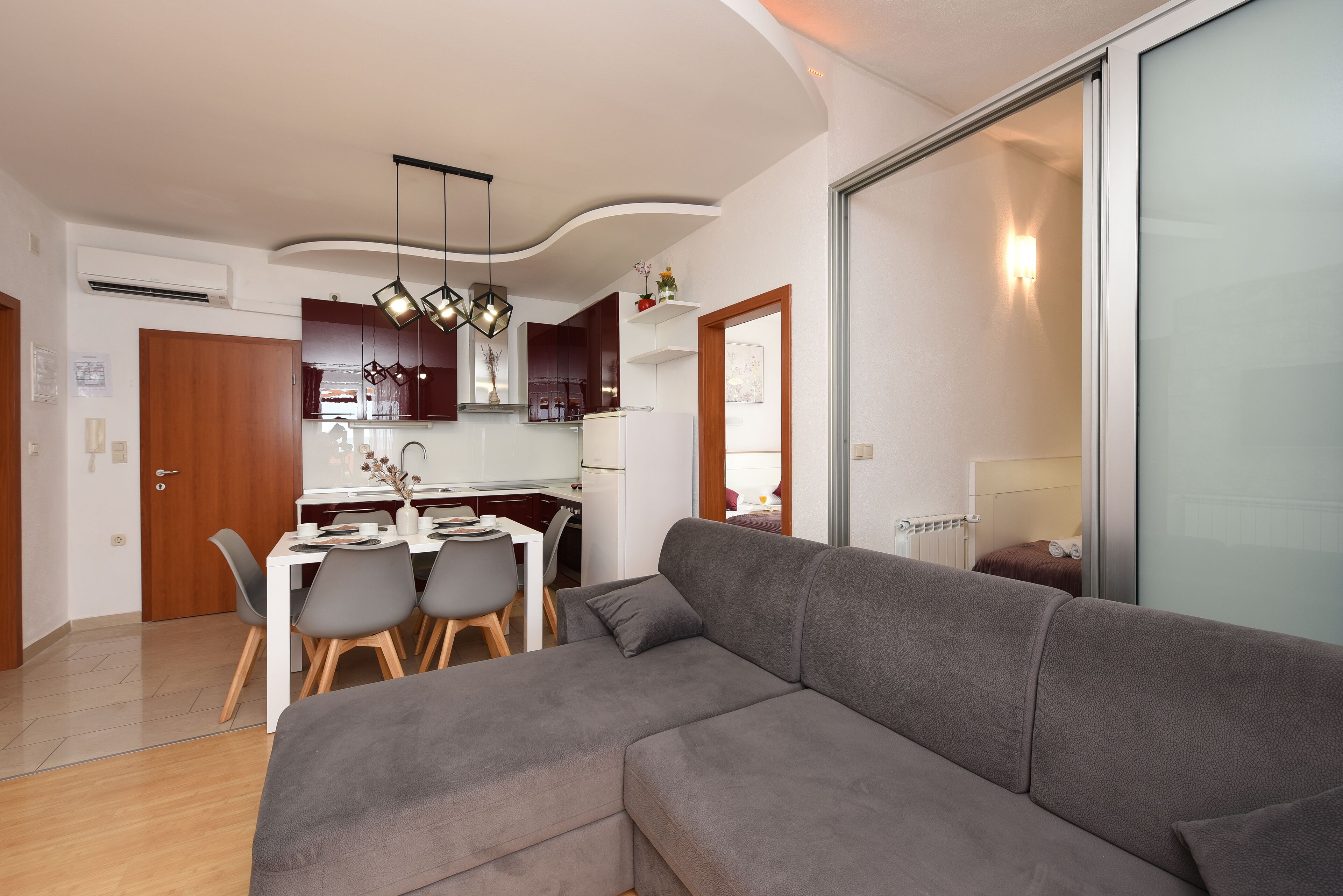 A modern living room with a grey sofa, a dining area with a white table and chairs, and a kitchen in the background.
