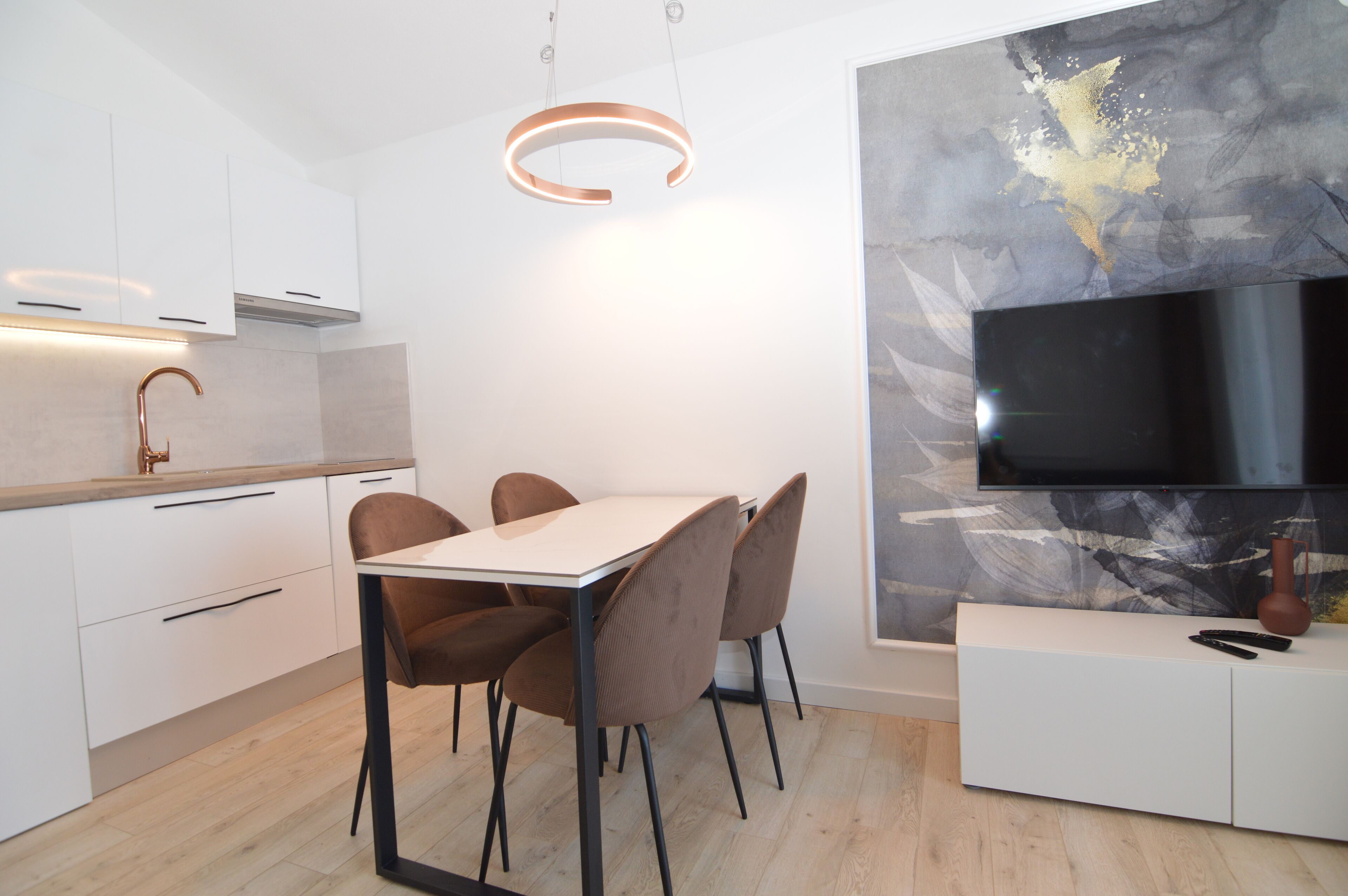 Une cuisine moderne avec des meubles blancs, un plan de travail en bois, une table à manger avec des chaises, un luminaire suspendu et un téléviseur à écran plat fixé au mur, le tout agrémenté d’une œuvre d’art évoquant le marbre.