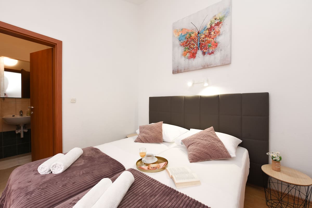 A neatly made bed with pillows, a bedside table with a book and a glass of orange juice, and a wall painting.