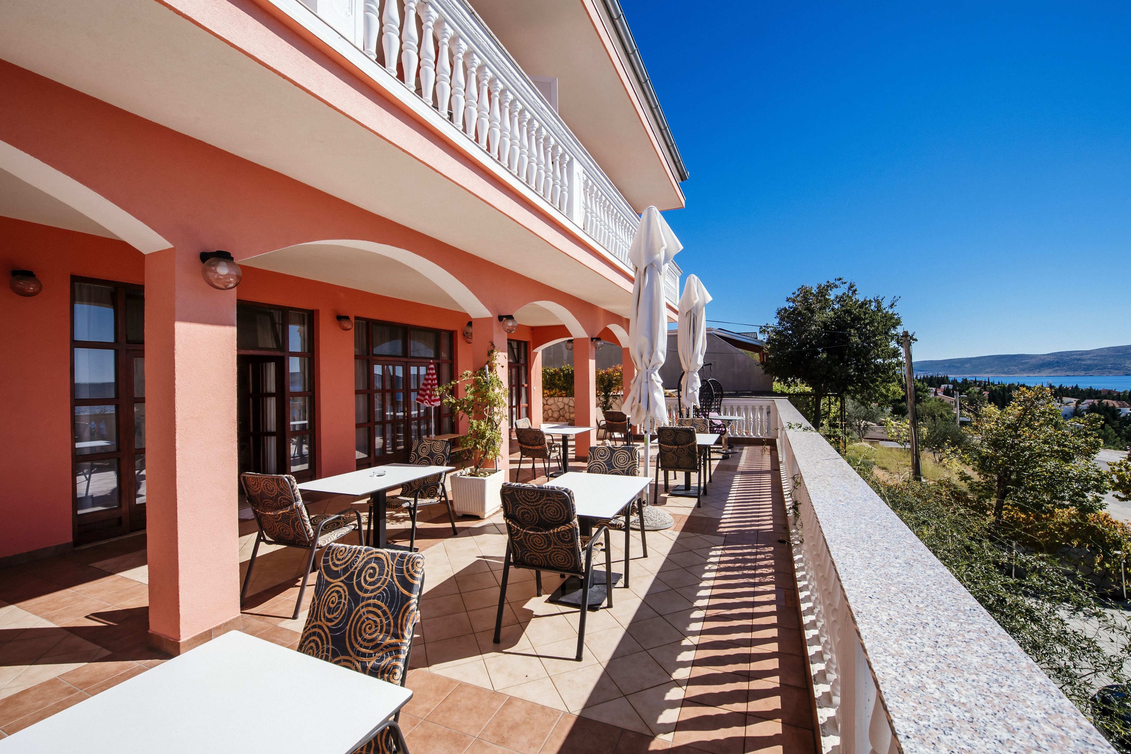An outdoor patio with tables and chairs, a balcony with white balustrades, and a view of the landscape.