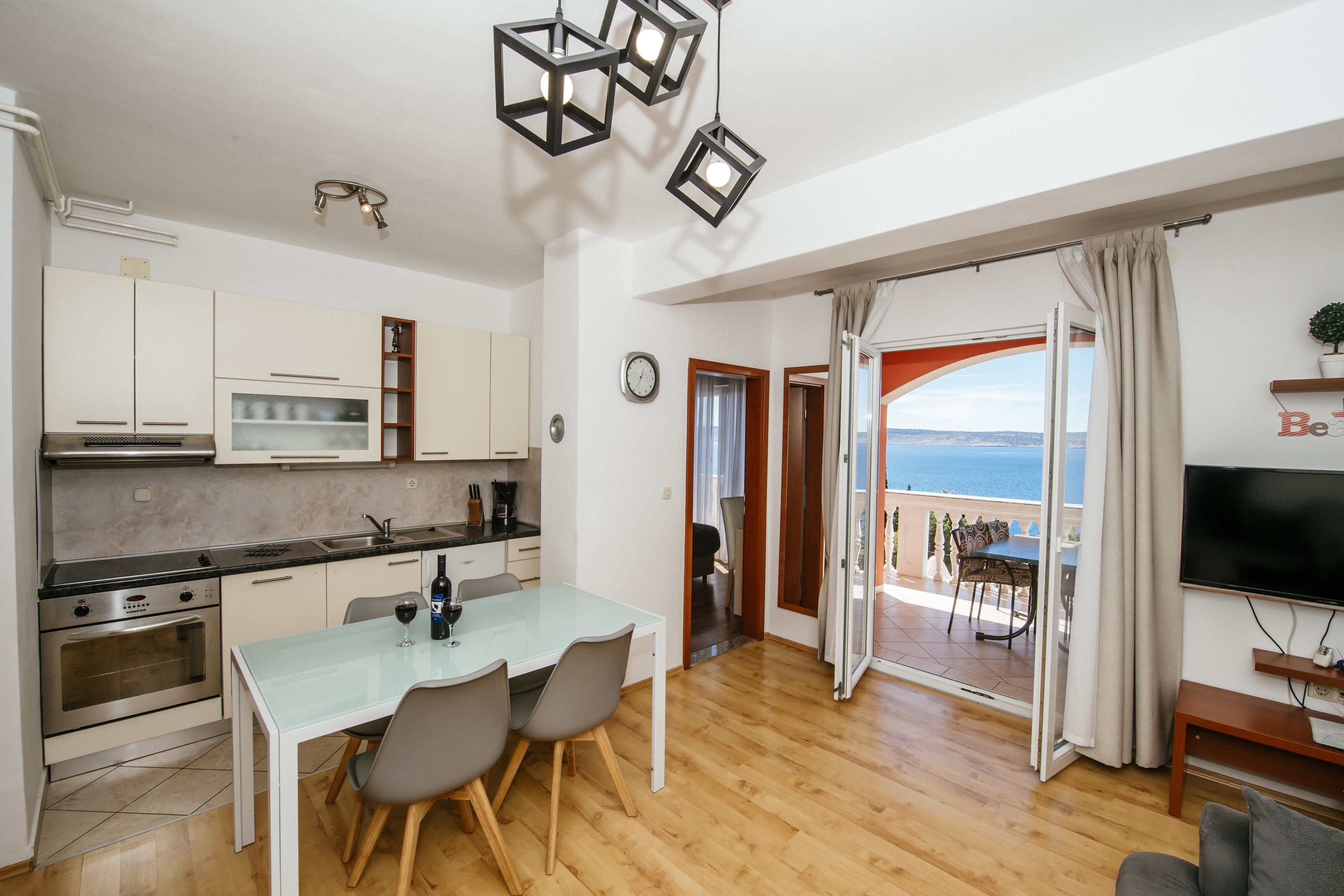 A modern kitchen with white cabinets, a dining table with chairs, a TV, and a balcony with a view of the sea.