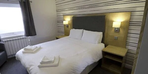 Habitación de hotel con una cama bien hecha, dos mesitas de noche con lámparas, una ventana con cortinas y un radiador.