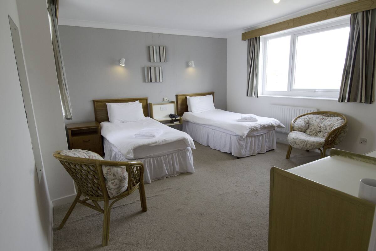 A hotel room with two single beds, a wooden chair, a desk, and a window with curtains.