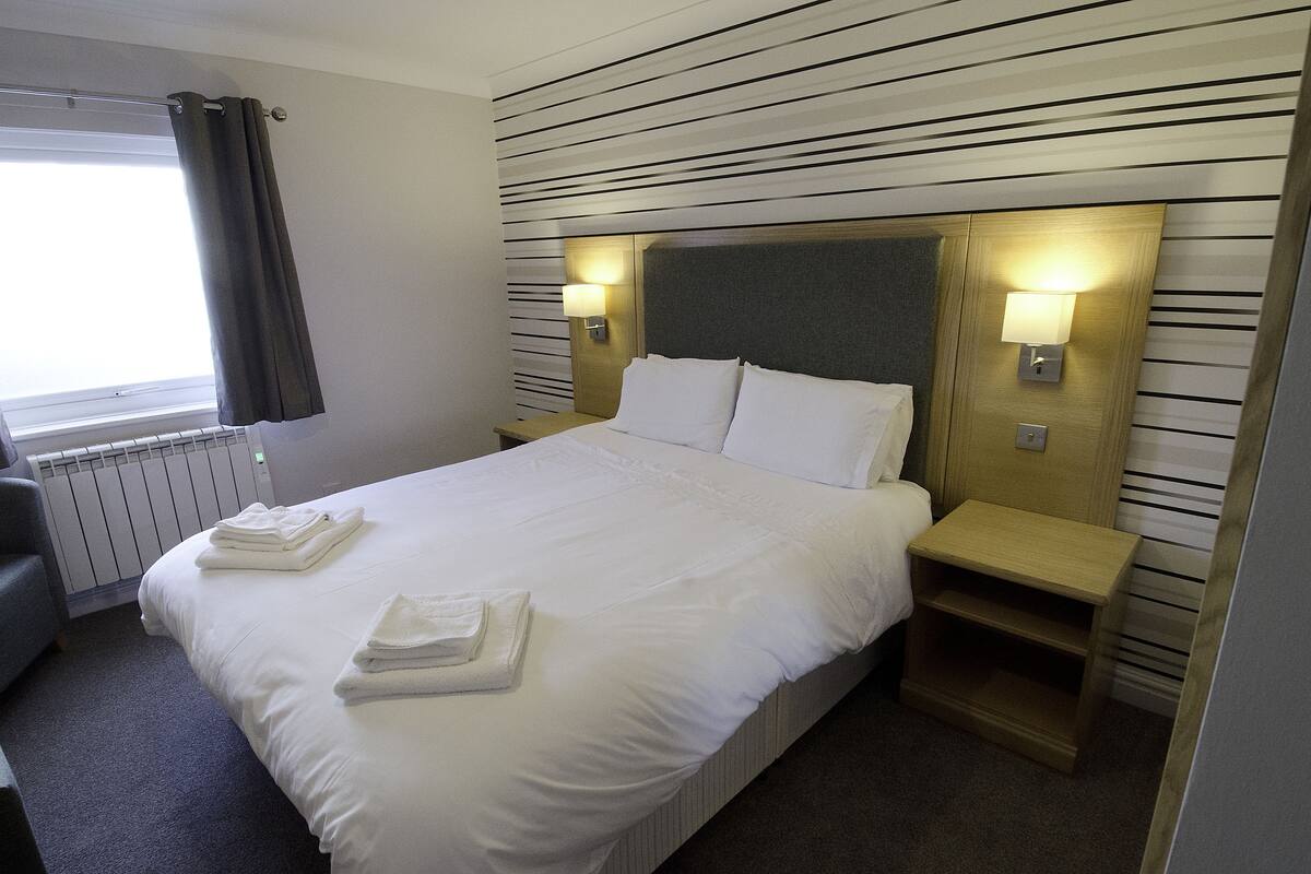 A neatly made bed with white linens and towels, a bedside table with a lamp, and a radiator.