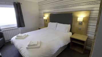 Een opgemaakt bed met witte lakens en handdoeken, een nachtkastje met een lamp en een radiator.