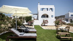 A sunlit outdoor lounge area with lounge chairs and a table, surrounded by a well-maintained lawn and a white building with blue accents. - Naxian Resort (Naxos)
