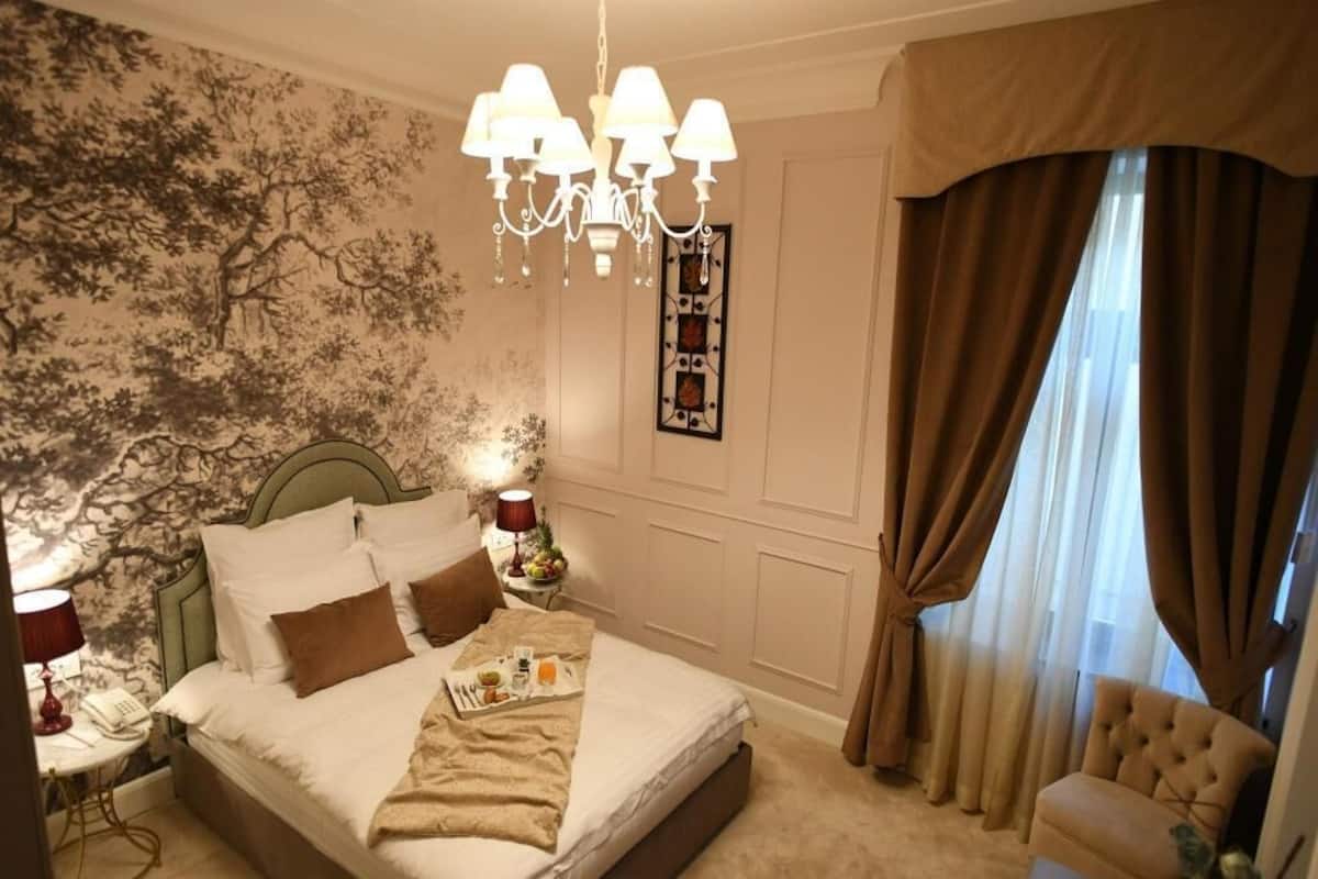 A hotel room with a bed, a chair, a chandelier, a nightstand, and a window with curtains.