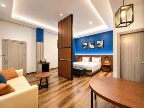 A modern hotel room with a bed, a sofa, a dining table, and a small kitchen area.