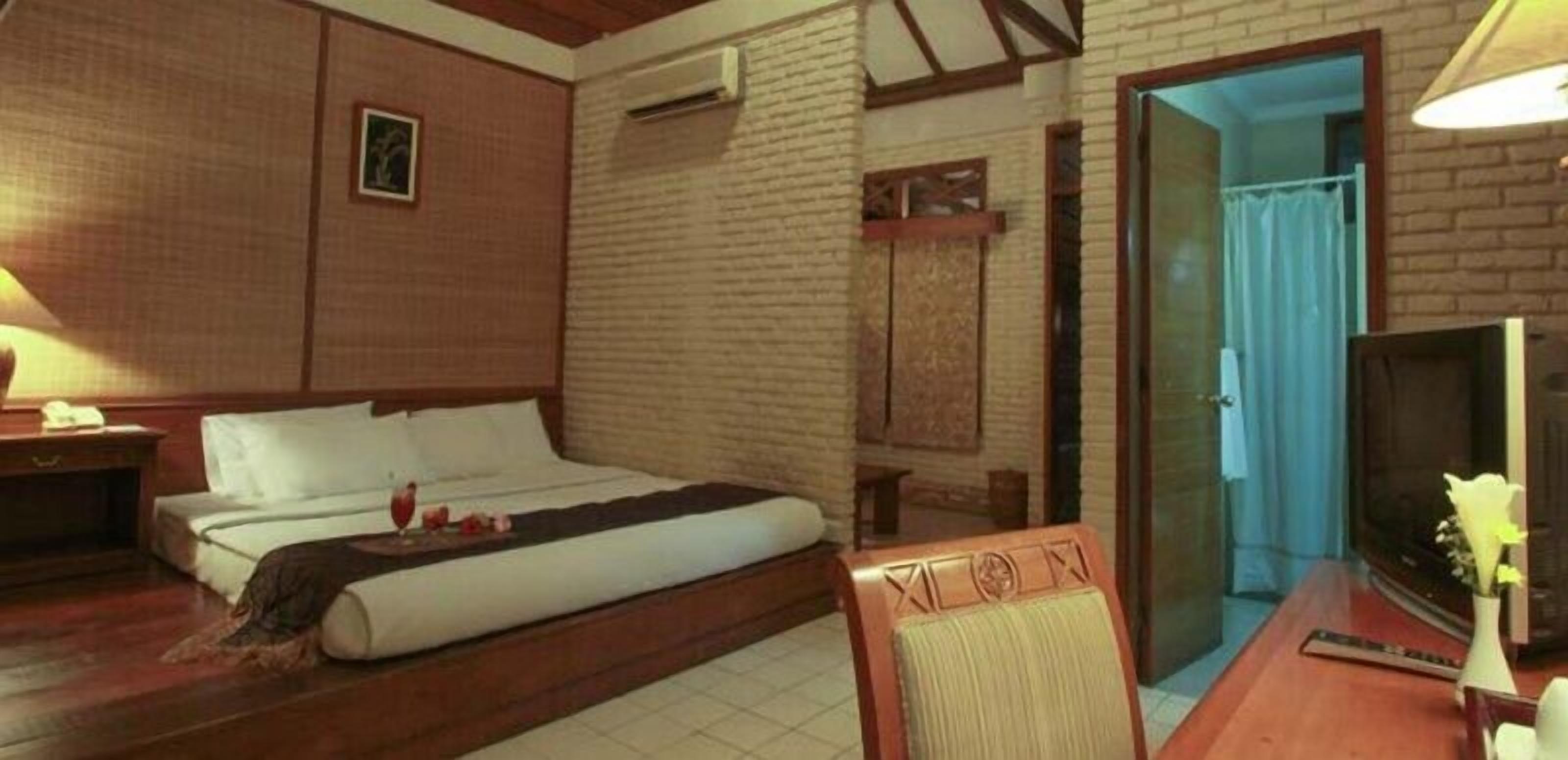 A hotel room with a bed, a wooden desk, a chair, a TV, and a door leading to another room.
