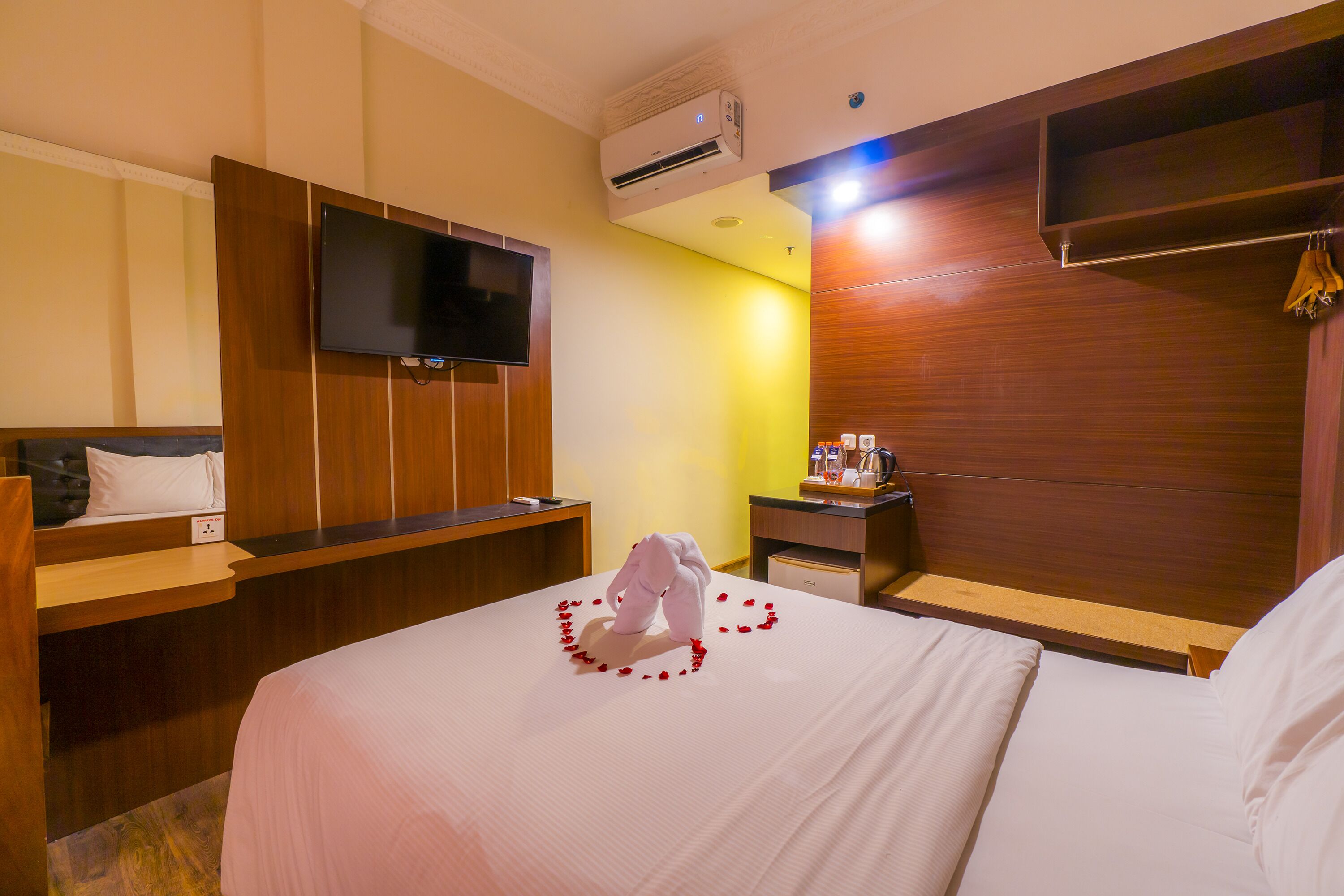 A hotel room with a bed, a TV mounted on the wall, a wooden headboard, a bedside table with a lamp, and an air conditioning unit.