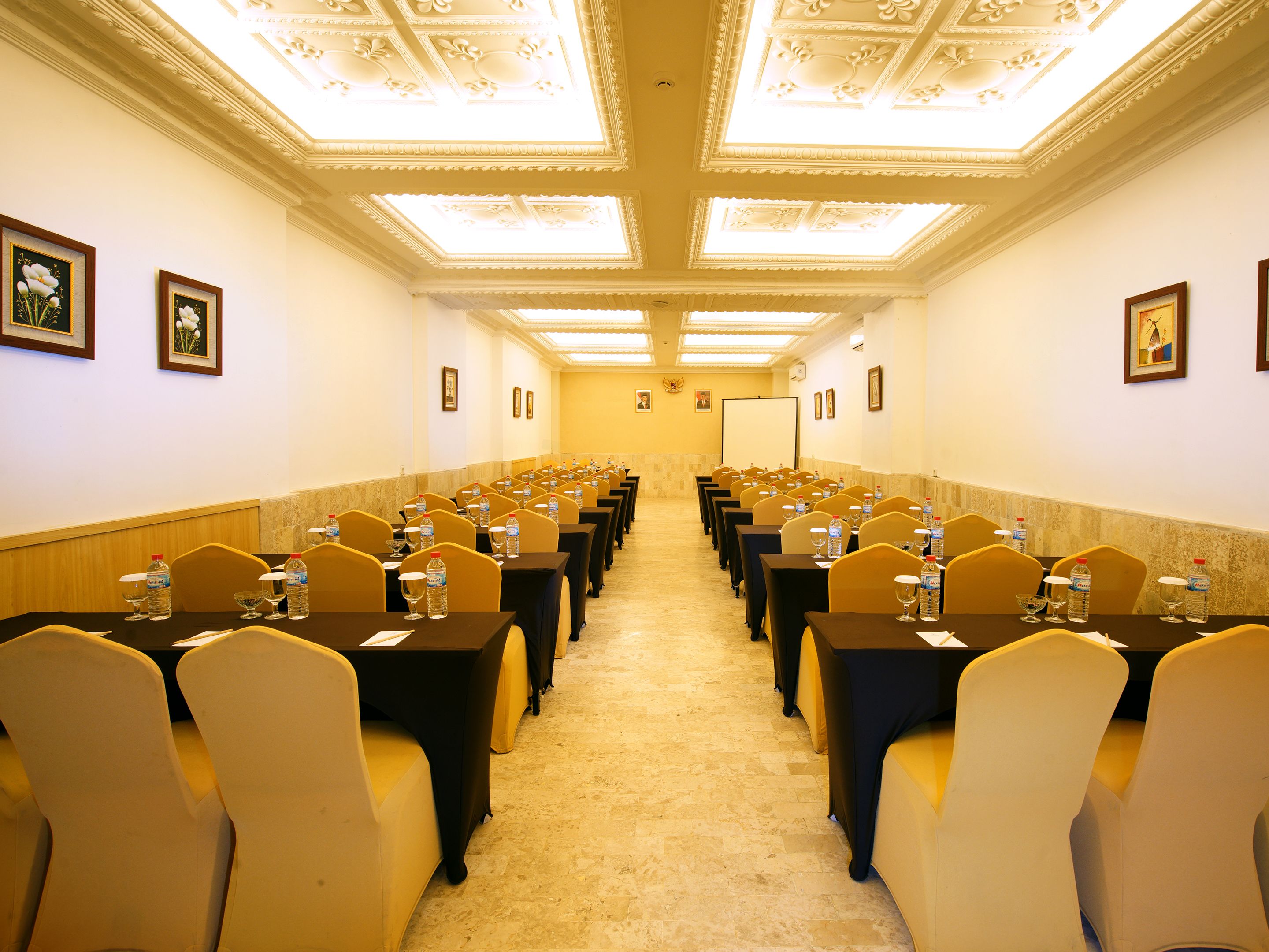 Une salle de conférence avec des rangées de tables et de chaises, équipée de bouteilles d’eau et de verres, et un grand écran à l’avant.