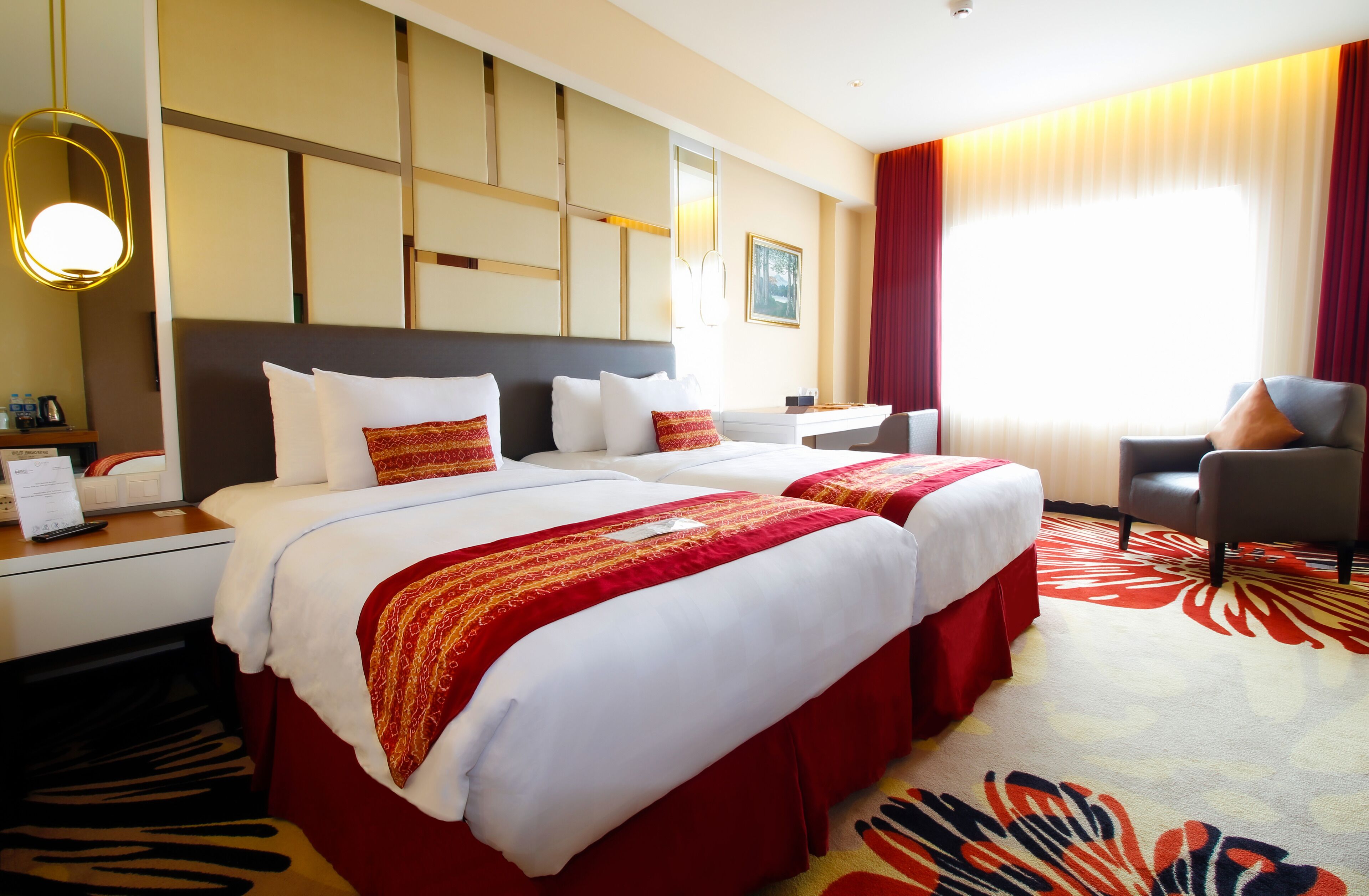 Quarto de hotel com duas camas, uma escrivaninha, uma cadeira e uma janela grande com cortinas.