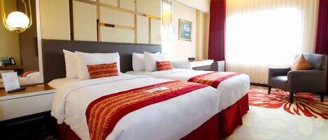 Quarto de hotel com duas camas, uma escrivaninha, uma cadeira e uma janela grande com cortinas.
