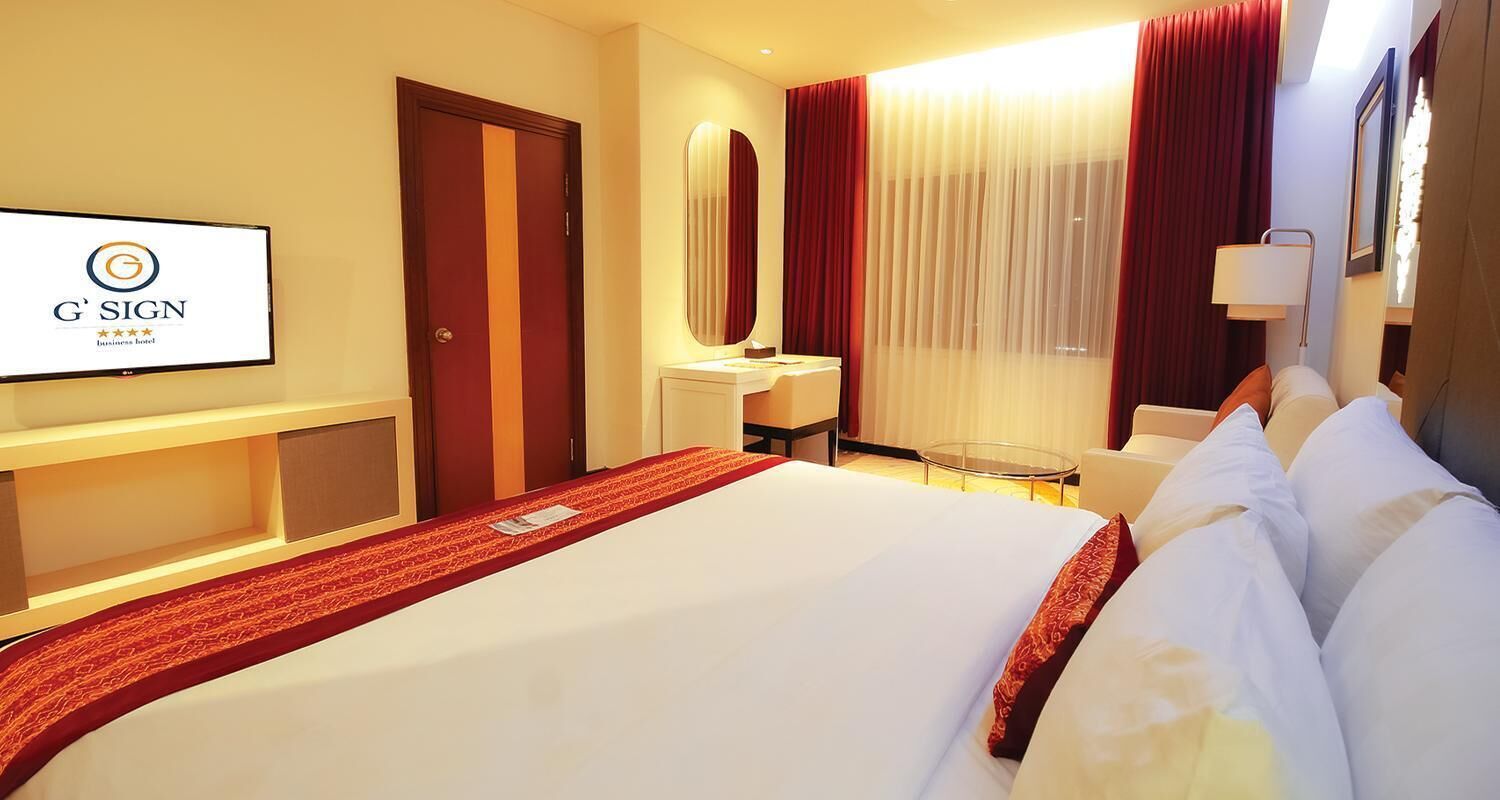 A hotel room with a bed, a desk, a chair, a mirror, and a television displaying the G'Sign logo.