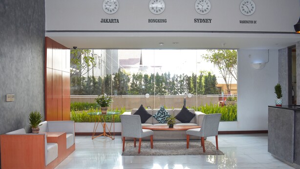 Un vestíbulo moderno con un conjunto de sofás, una mesita de café y un mostrador de recepción. Se observan relojes que marcan la hora en diferentes ciudades, y una vista de vegetación al exterior.