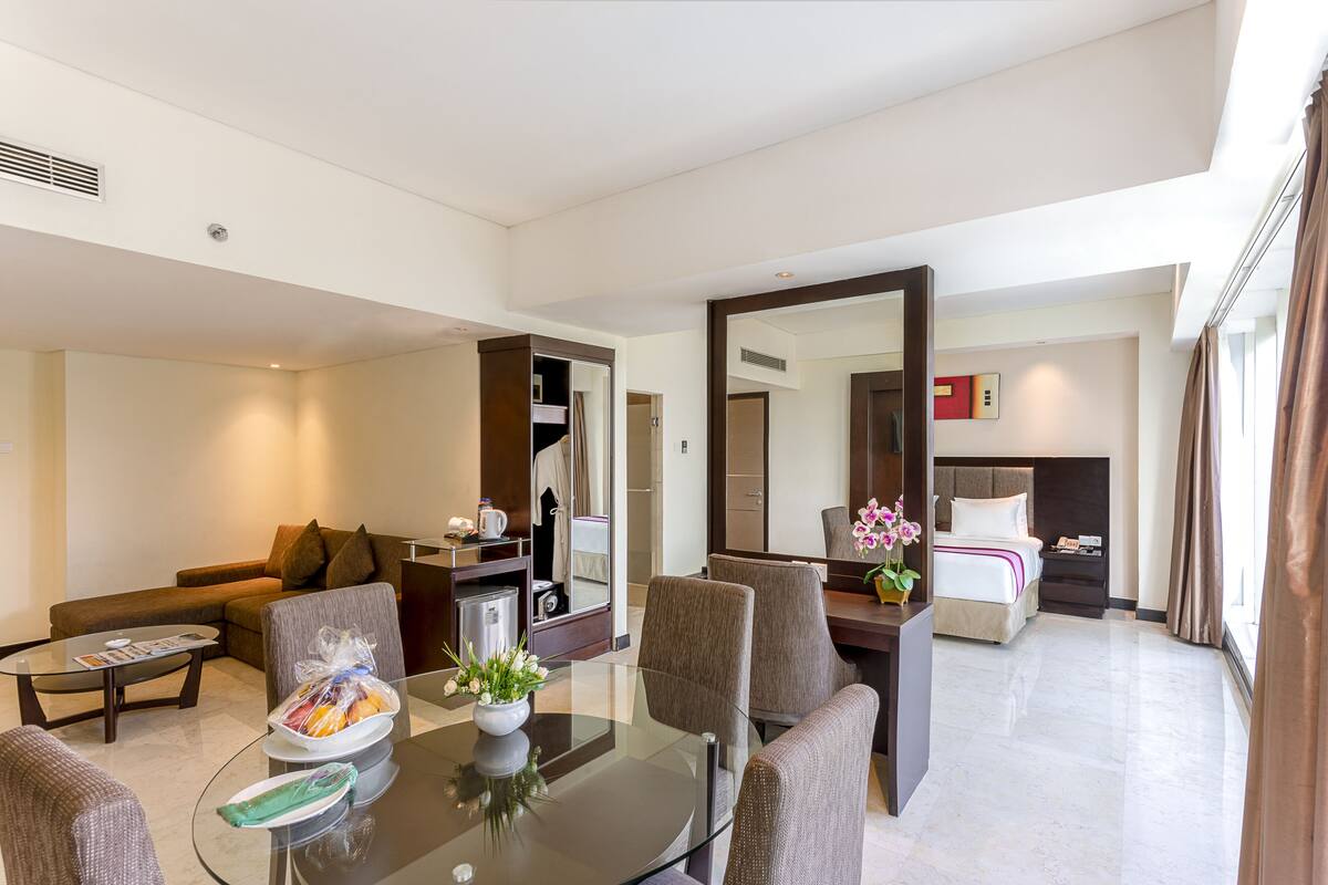 A modern hotel room with a glass dining table, comfortable seating, a large mirror, a neatly made bed, and a view of the city.