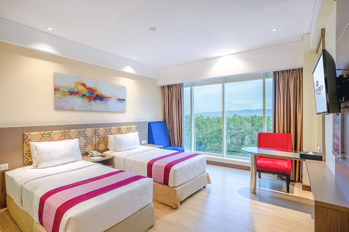 A hotel room with two beds, a desk, a chair, a TV, and a large window with a view.