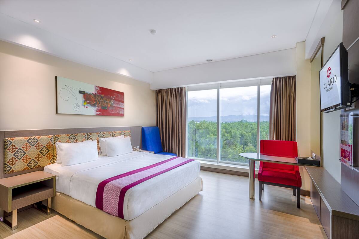A hotel room with a large bed, a desk, a red chair, a TV, and a window with a view of the outdoors.