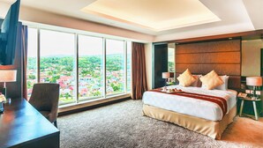 A hotel room with a large bed, a desk, a chair, a TV, and a view of a cityscape through large windows. - Claro Kendari (Kendari)