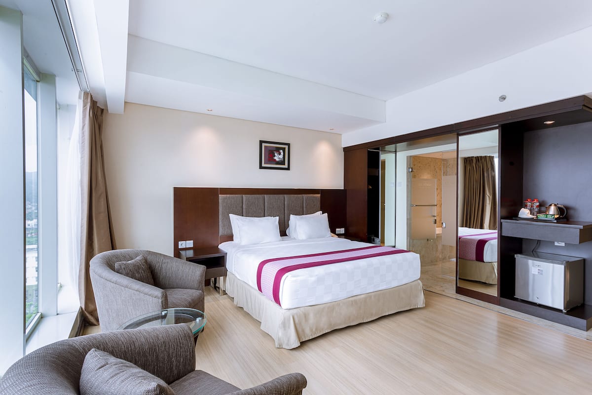 A modern hotel room with a large bed, a sitting area with a sofa and armchair, a small table, and a minibar.