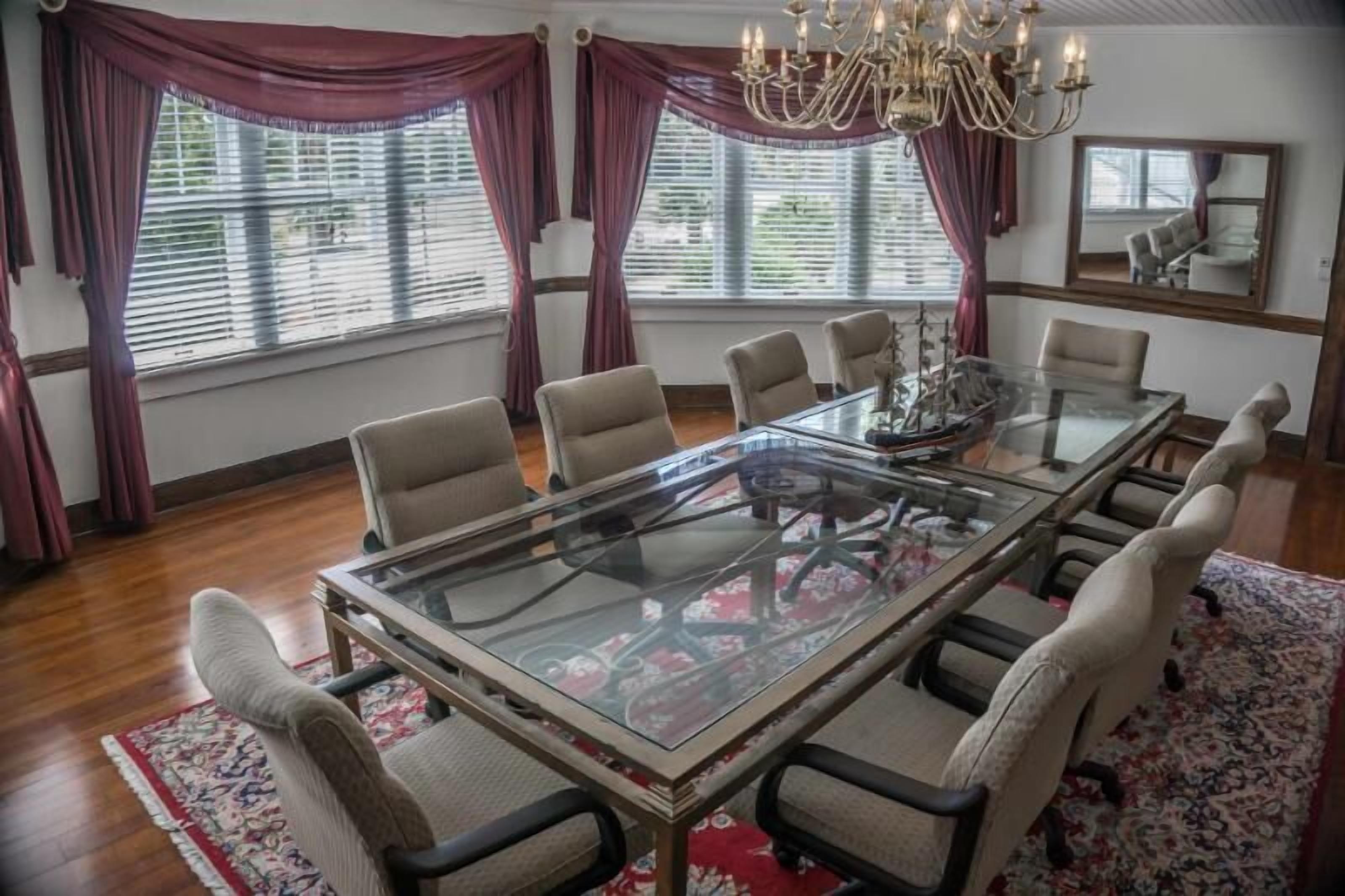 A conference room with a glass table, beige chairs, a chandelier, and large windows with curtains.