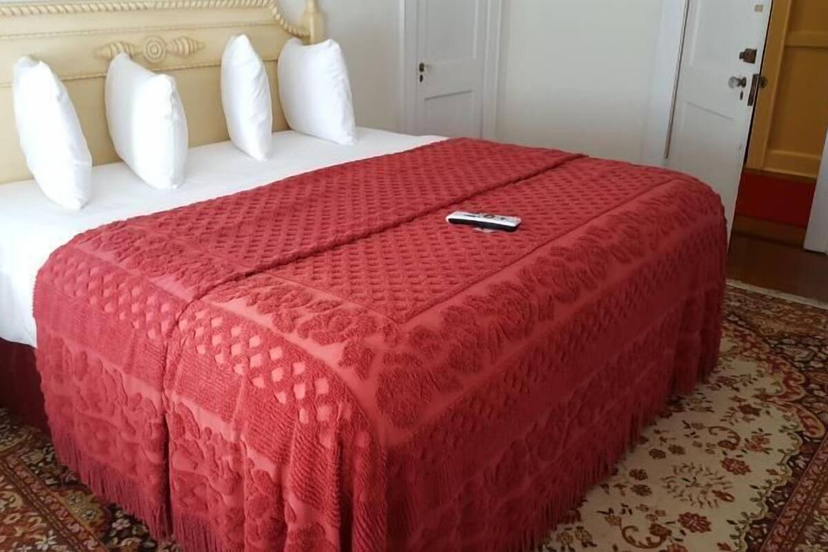 Un lit bien fait, recouvert d’une couvre-lit rouge à motifs et agrémenté d’oreillers blancs, une télécommande posée sur le lit et un tapis à motifs sur le sol.