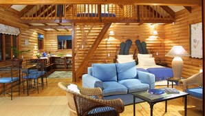Interior de una cabaña de troncos con una escalera de madera, un sofá azul, sillas de mimbre y un comedor.