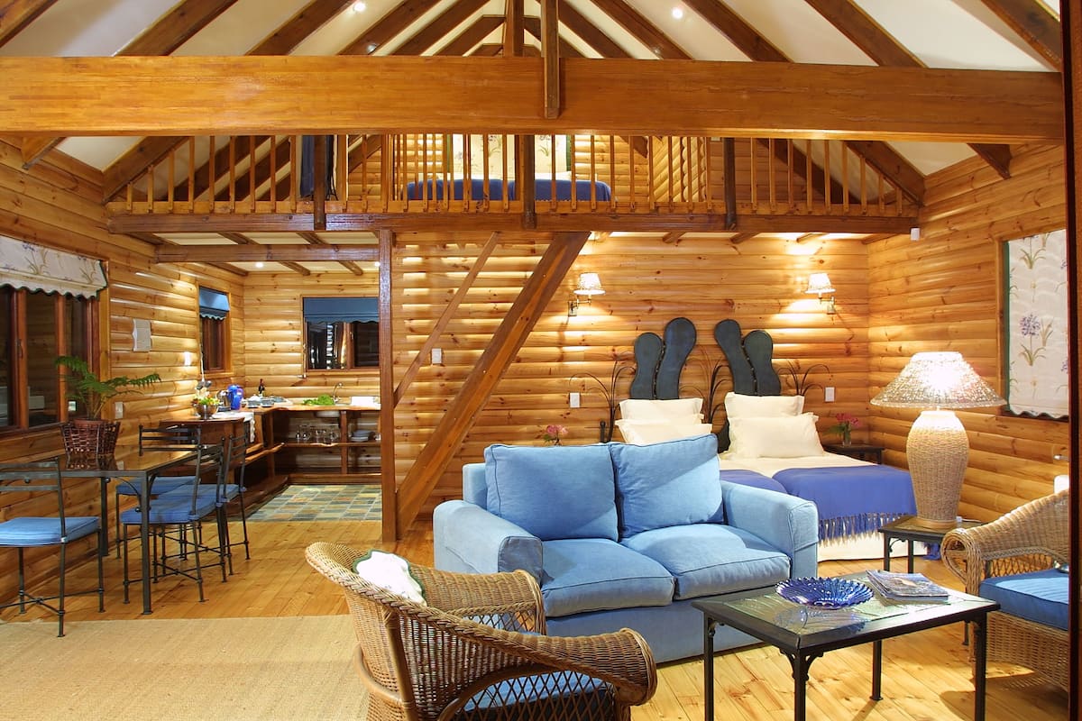 A log cabin interior with a wooden staircase, a blue sofa, wicker chairs, and a dining area.