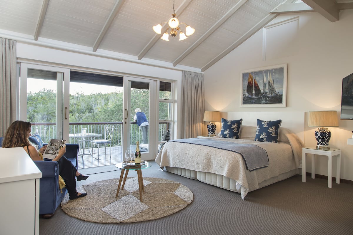 A bedroom with a large bed, a sitting area with a sofa and a table, and a balcony with a view of the outdoors.