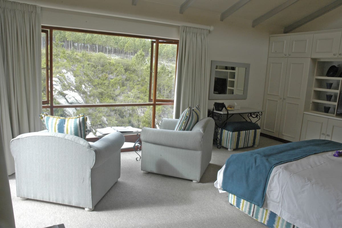 A bedroom with a large window, a bed, a chair, a small table, and a wardrobe.