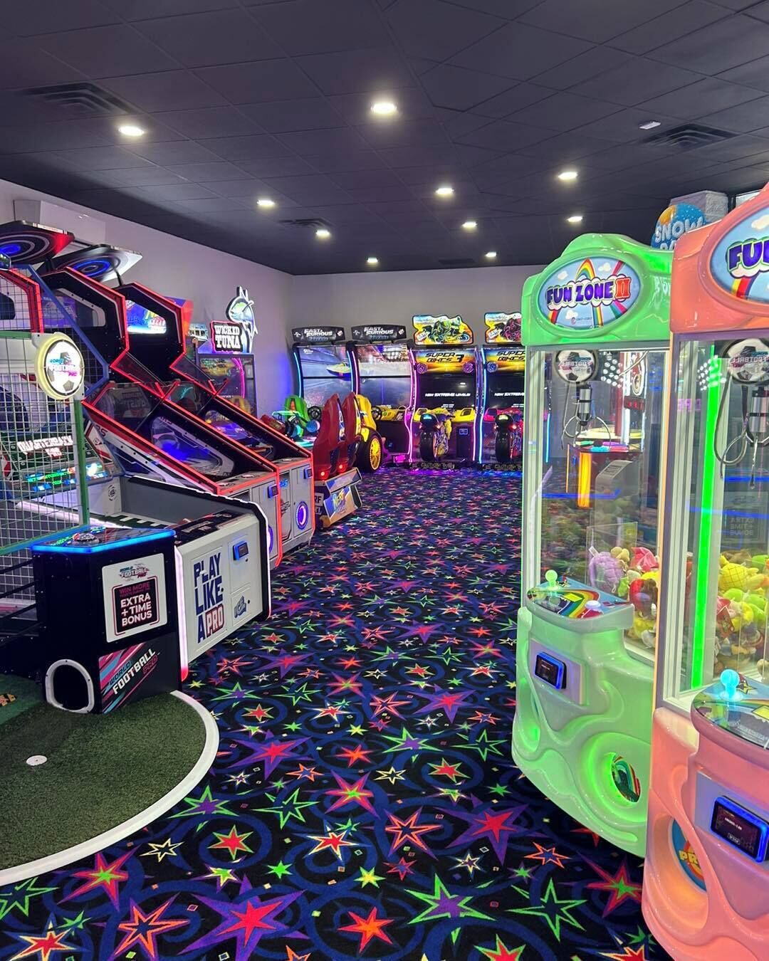 Un salón de juegos interior con varias máquinas recreativas, una alfombra estampada e iluminación empotrada en el techo.