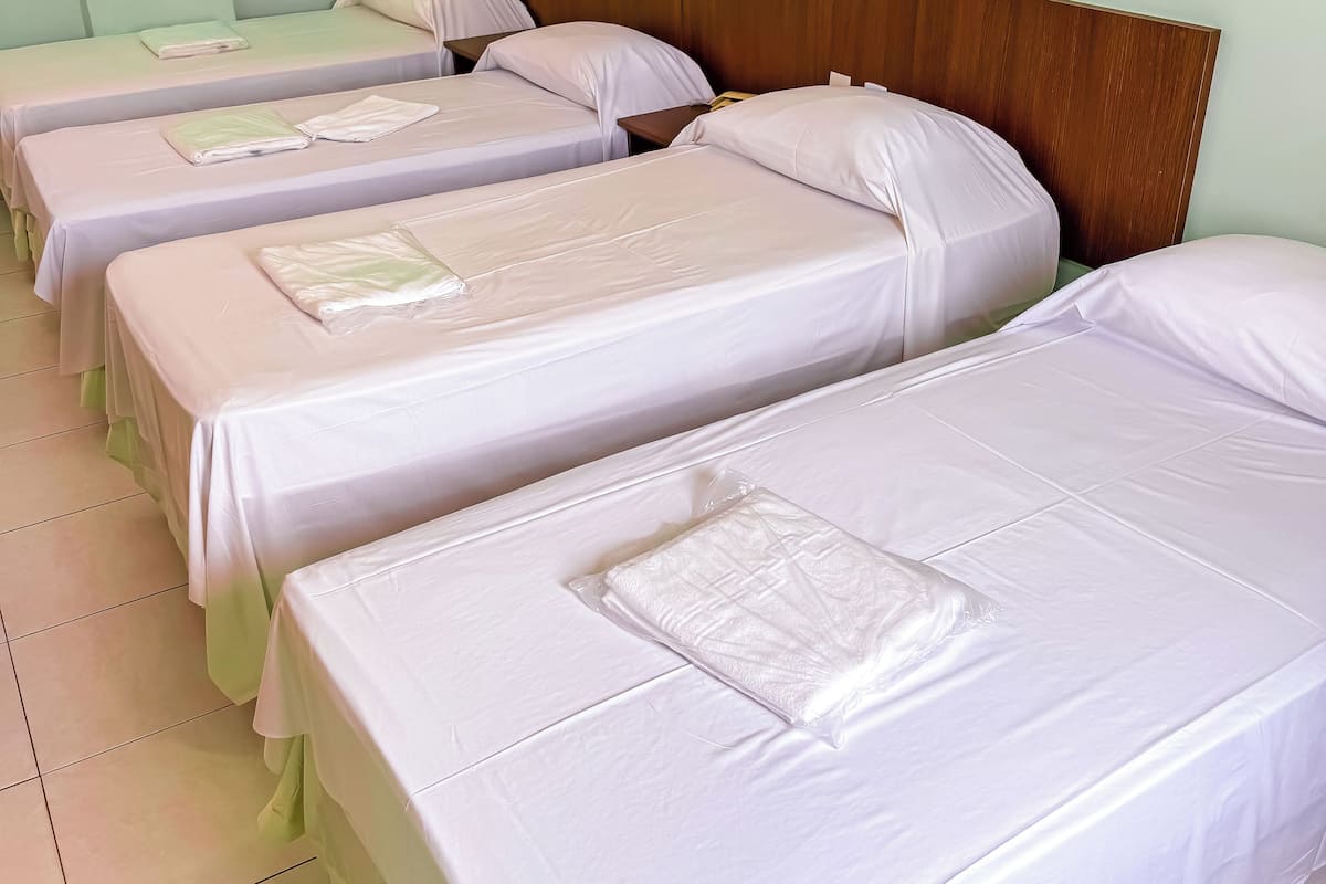 A row of neatly made beds with white linens and pillows, wooden headboards, and a tiled floor.