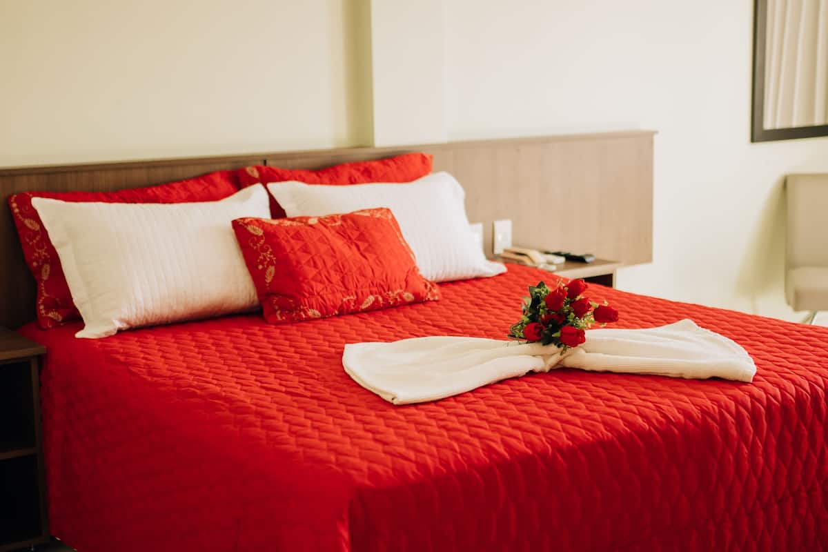 A hotel room with a red quilted bedspread, white pillows, and a bouquet of red roses.