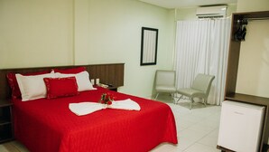 A hotel room with a large bed, a small desk, a chair, a refrigerator, and a window with curtains. - Trindade Plaza Hotel (Trindade)