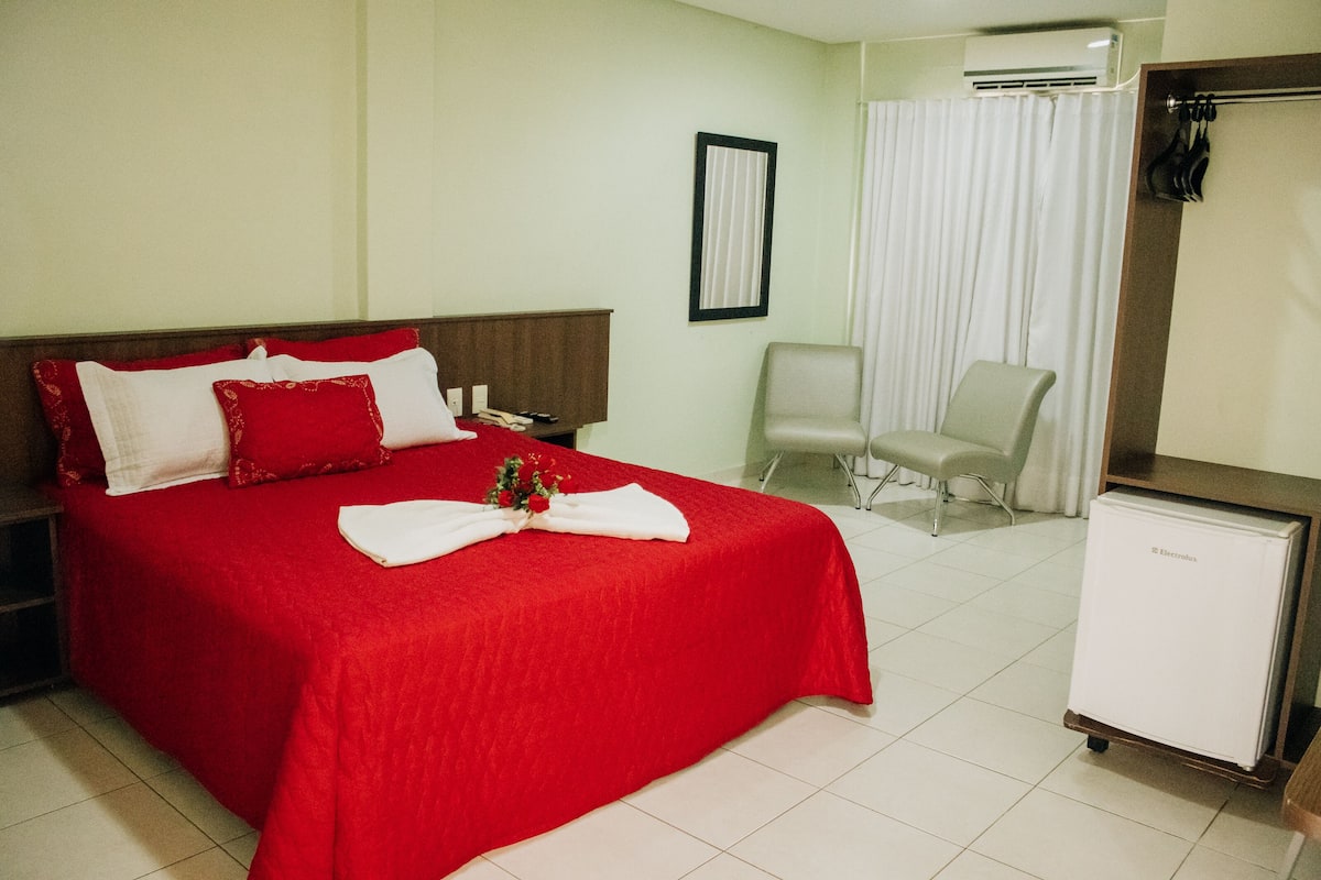 A hotel room with a large bed, a small desk, a chair, a refrigerator, and a window with curtains.