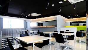A modern dining area with white tables and black chairs, a kitchen area with green accents, and a staircase leading to an upper level. - Swift Inn Aeropolis Airport (Tangerang)