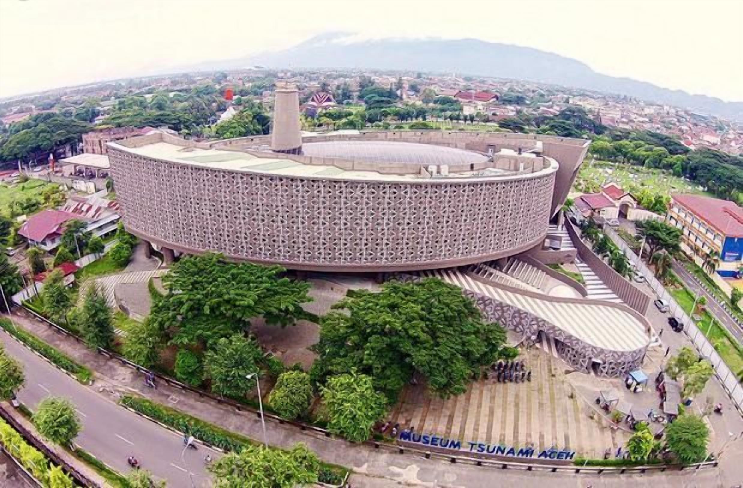 A large, circular building with a patterned exterior, surrounded by greenery and a road.