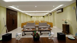 Ein Konferenzraum mit Bühne, Reihen von Stühlen, zwei Sofas, Tischen mit Wasserflaschen und einem Topf mit Pflanze.