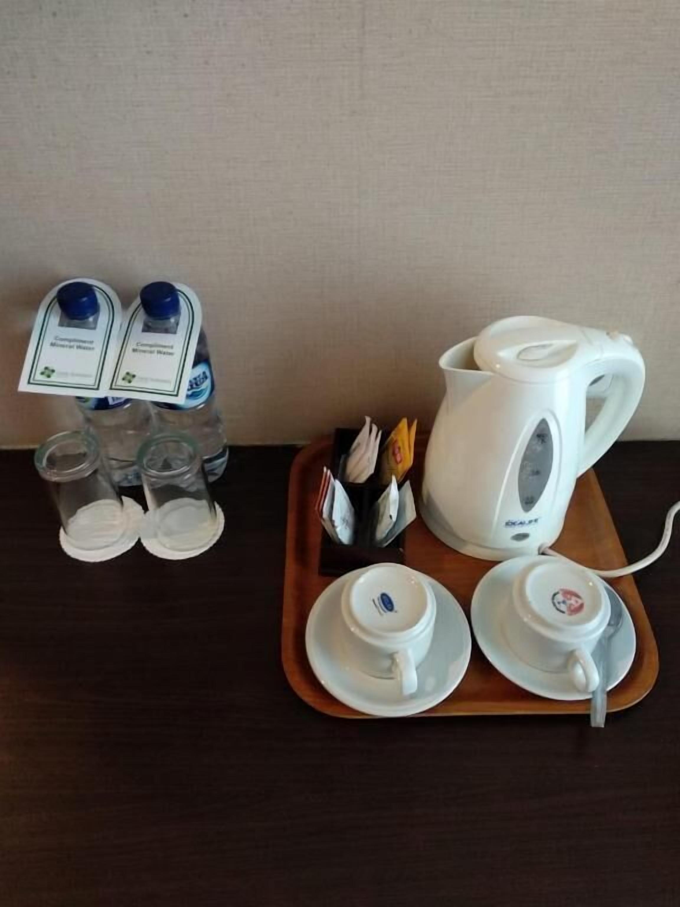 A hotel room's breakfast setup with a white electric kettle, two cups, and a tray with a mug and utensils.