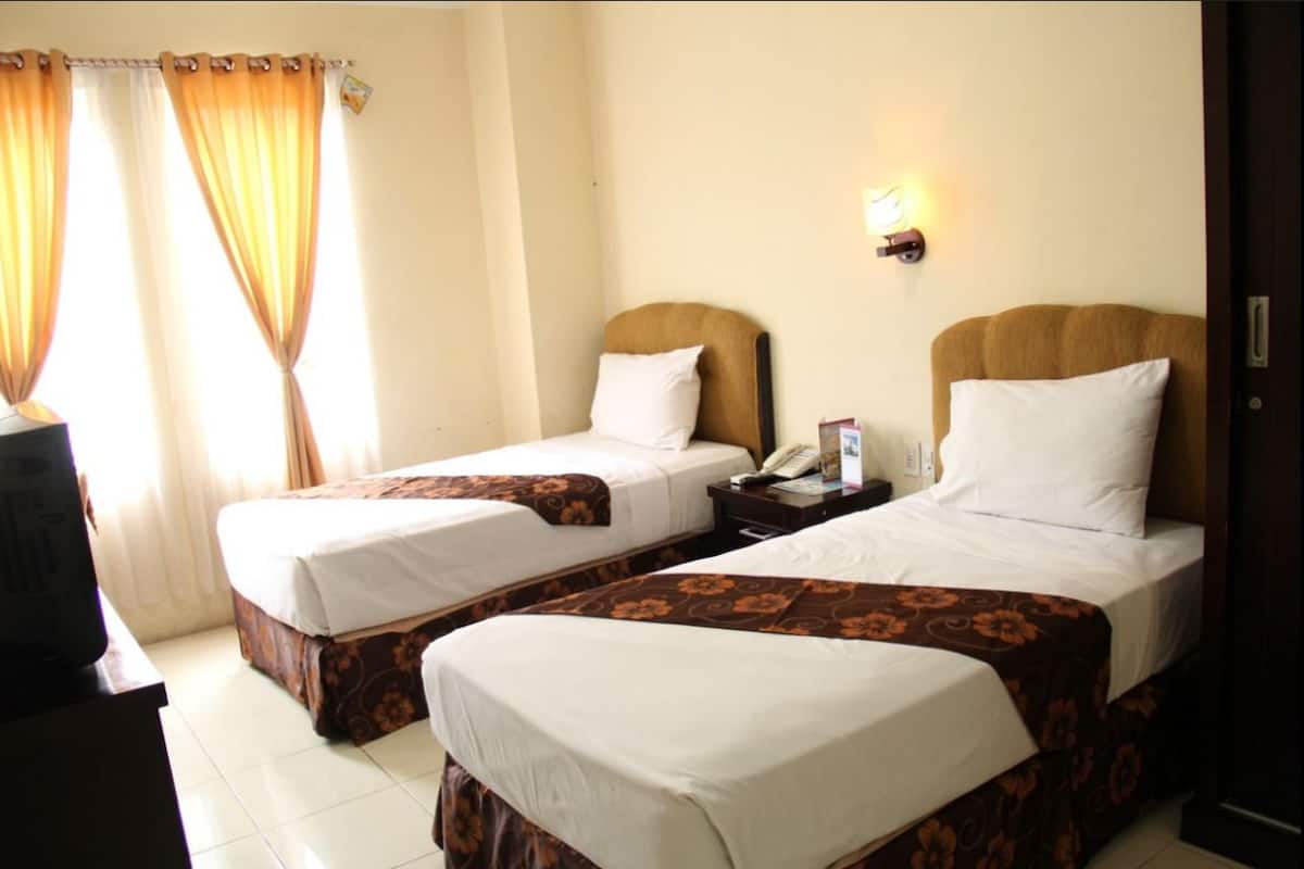 Una habitación de hotel con dos camas individuales, una mesita de noche con lámpara y una ventana con cortinas.
