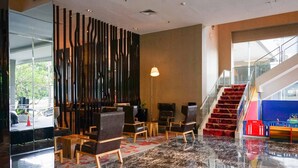 A lobby with a modern design, featuring a marble floor, a staircase with a red carpet, and a seating area with chairs and tables. - Premiere by Grand Zuri (Pekanbaru)