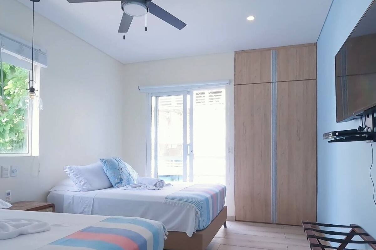 A bedroom with two beds, a ceiling fan, a window with blinds, a wardrobe, and a TV mounted on the wall.