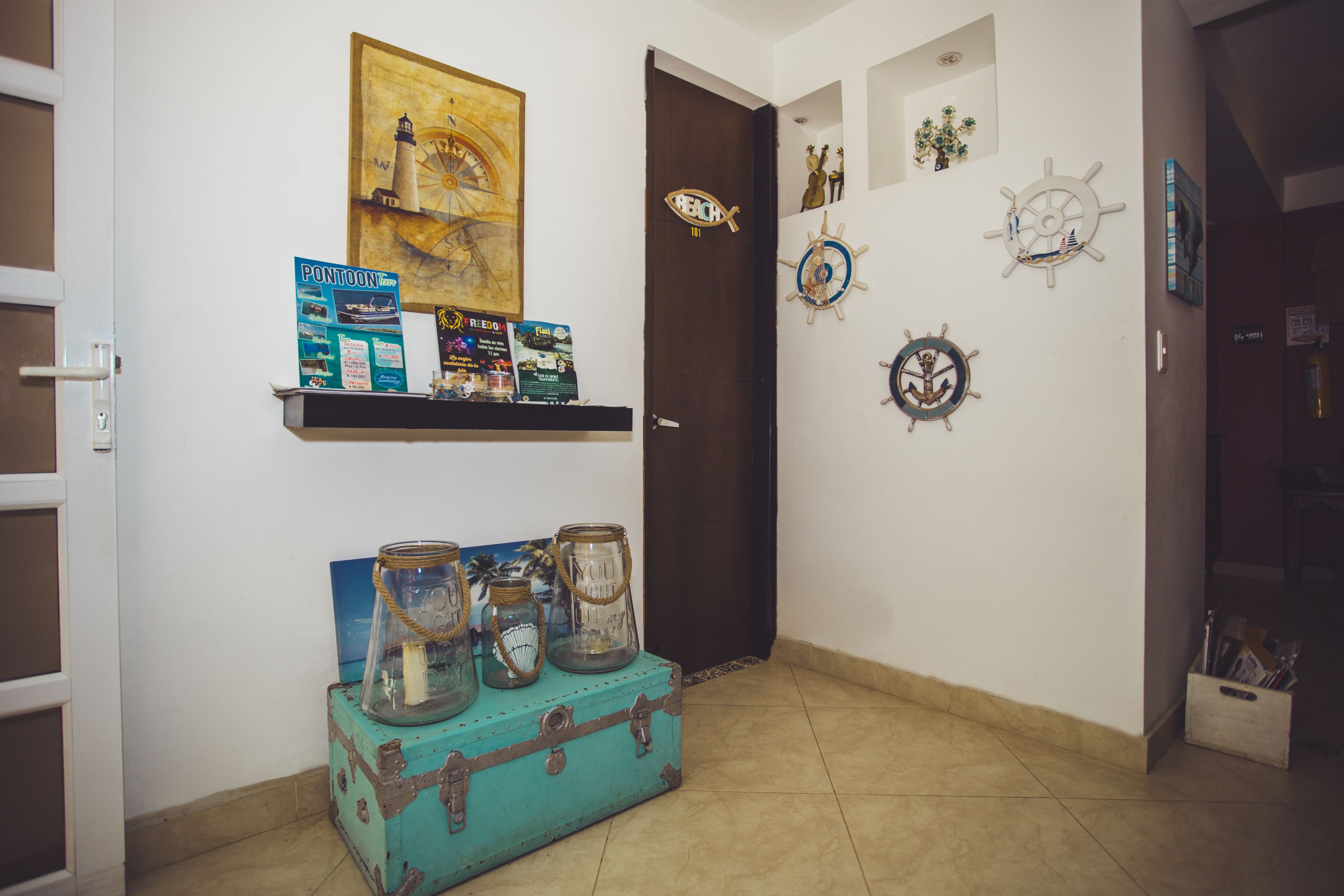A room with a wooden door, a wall-mounted shelf with travel-themed items, a blue suitcase, and nautical decorations.