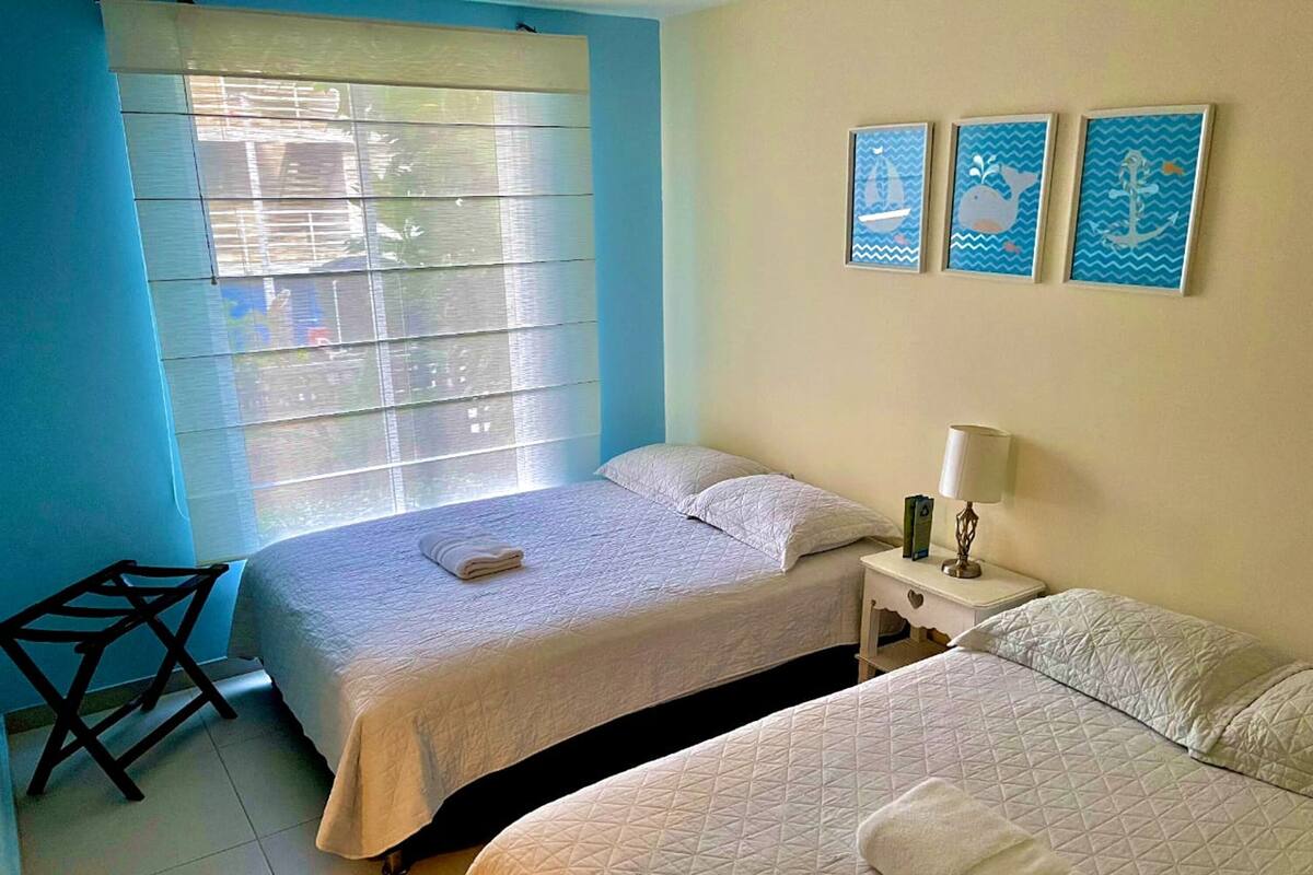 A hotel room with two beds, a nightstand, a chair, and a window with blinds.